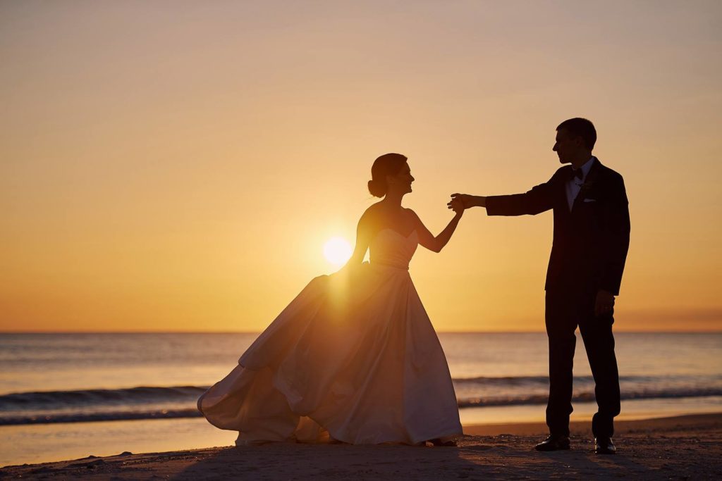 A bride and groom are depicted in silhouette. They are on a beach and it is sunset. 