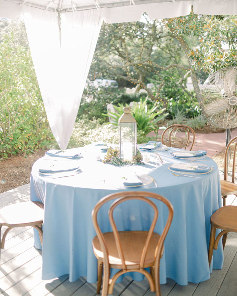 A table setting at a wedding is displayed. The table covering is a light blue and the chairs are tan with a rounded back. A glass lantern completes the centerpiece.