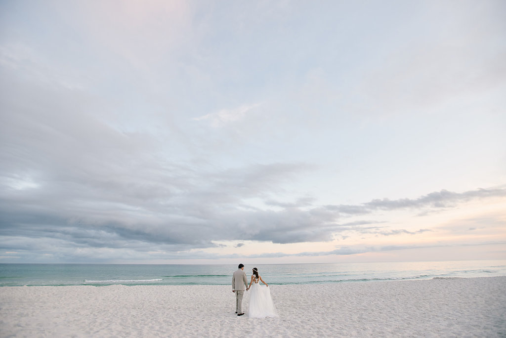 A bride and groom are facing away from the camera towards a beach. It is dusk. The bride and groom are wearing their wedding clothes in this portrait.