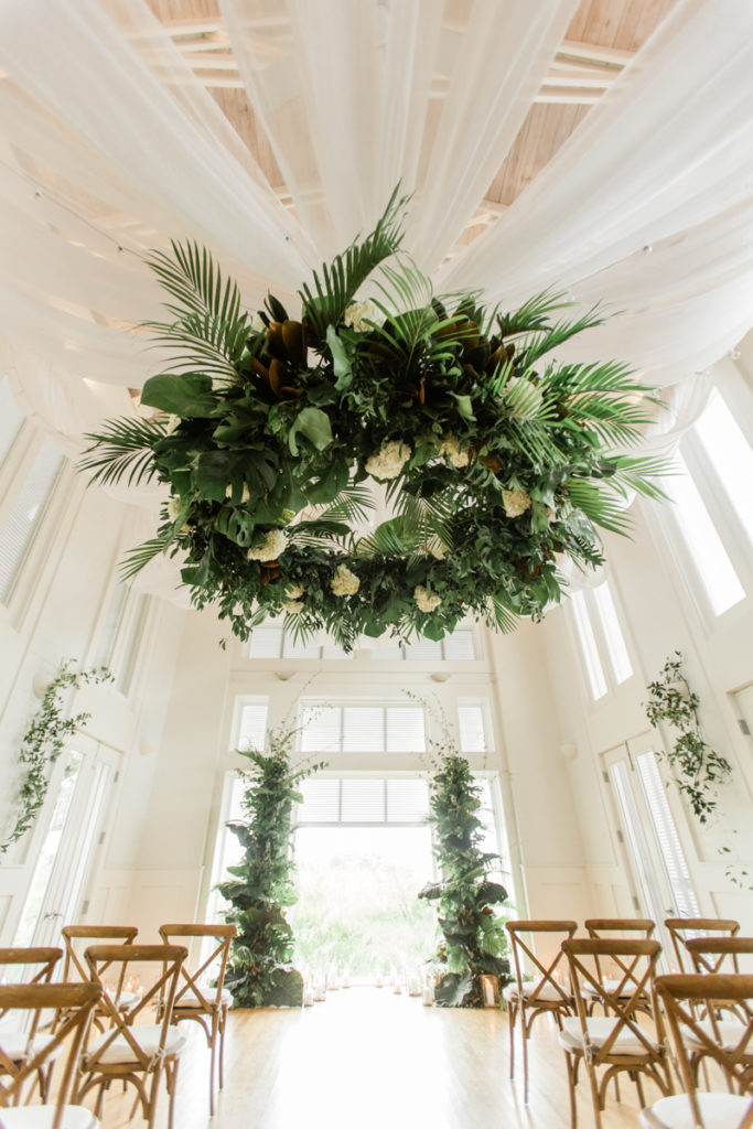 The Meeting House at Carillon Beach in 30A, Florida s decorated with light brown wooden chairs and a tropical altar and floral chandelier. The meeting house is a chapel-like room with wood floors and white walls and ceiling. 