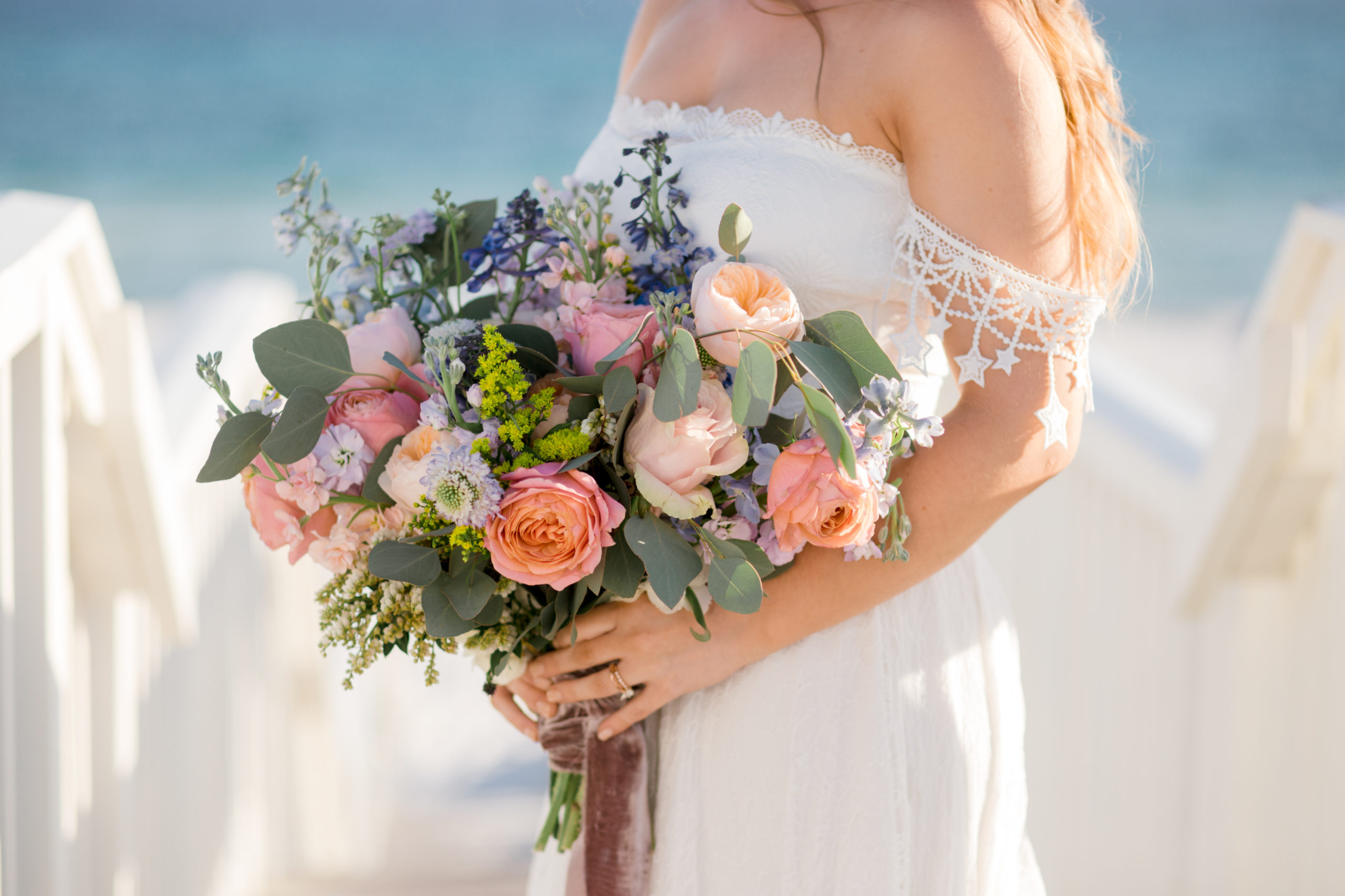 A bride stands by the ocean holding a bright bouquet of flowers.